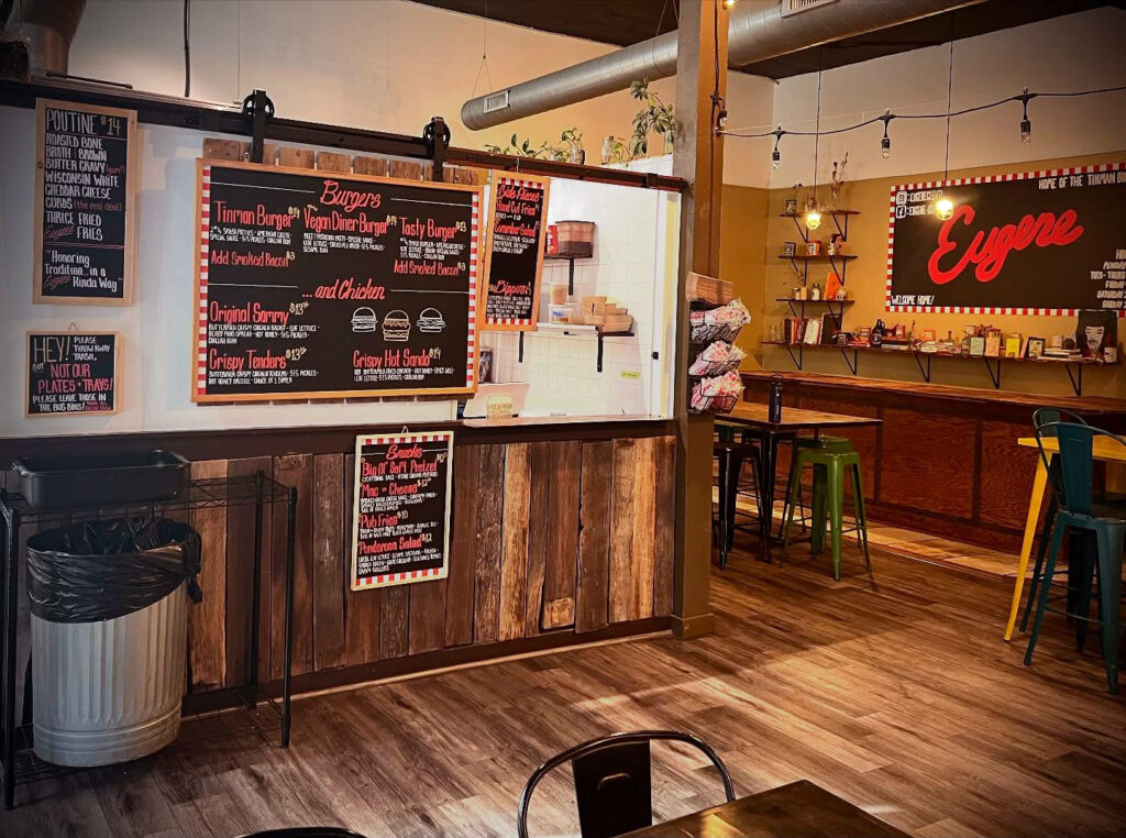 Eugene restaurant interior, chalkboard sign on the left with menu items, kitchen window, on the right large Eugene sign. Wood paneling and wood tables with metal bar stools.