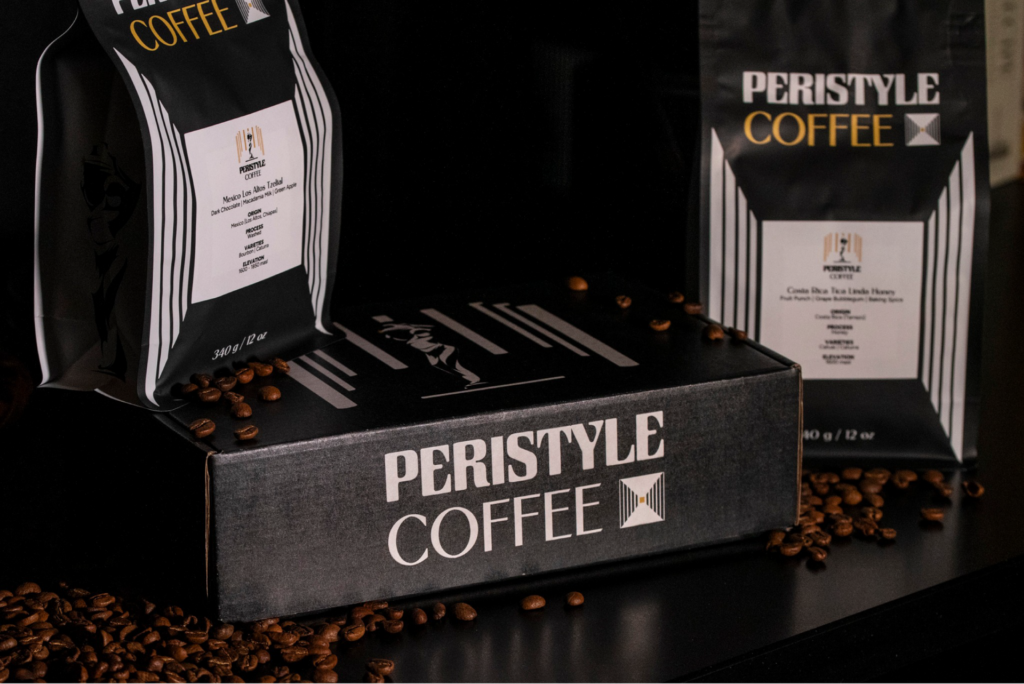 Product photo, white and gold bags and box in foreground, on a dark table surrounded by coffee beans